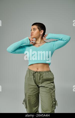 Eine junge Frau mit kurzen Haaren posiert selbstbewusst in trendiger Kleidung, einem Crop-Top und einer Cargo-Hose vor grauem Hintergrund. Stockfoto