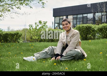 Eine junge Frau mit kurzen Haaren sitzt auf üppig grünem Gras vor einem modernen Gebäude. Stockfoto