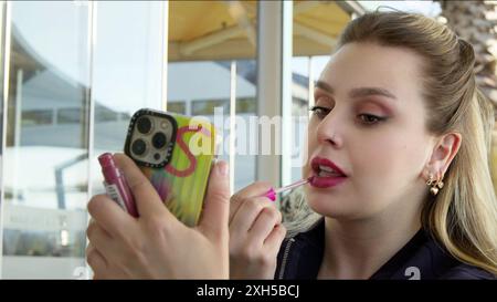 Die junge blonde Frau schminkt sich und benutzt das Handy als Spiegel Stockfoto