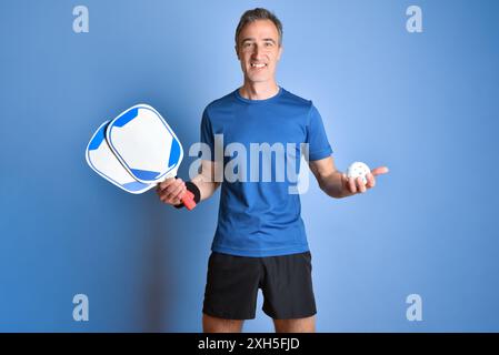 Lächelnder Mann in Sportbekleidung, der Pickleball-Paddel und Ball in den Händen auf blauem isoliertem Hintergrund zeigt. Stockfoto