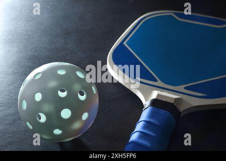 Blauer hölzerner Paddle-Pickleball-Schläger und ein weißer Ball auf schwarzem Tisch. Erhöhte Aussicht. Stockfoto