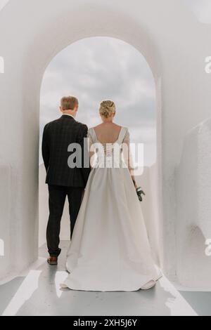 Eine Braut und ein Bräutigam spazieren an einem sonnigen Tag durch einen weißen Torbogen. Die Braut trägt ein weißes Kleid und der Bräutigam einen Anzug. Der Torbogen Stockfoto