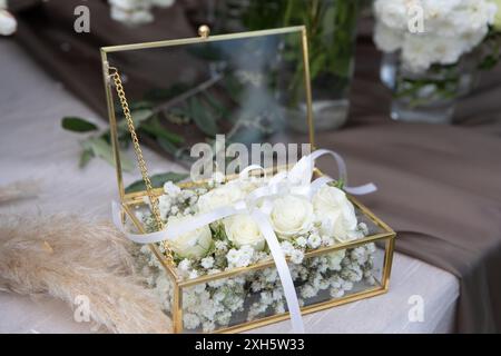 Elegante Glasschmuckdose gefüllt mit weißen Rosen und Babyatme, mit weißem Band gebunden, auf Tisch mit brauner Tischdecke gelegt Stockfoto