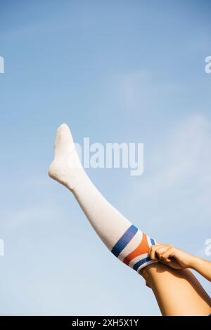 Junge Frau, die ihr Bein zum Himmel streckt und eine lange weiße Socke mit bunten Streifen anzieht. Stockfoto