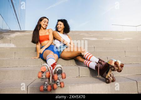 Zwei junge Frauen sitzen auf Stufen, entspannen und lächeln mit ihren Rollschuhen und genießen einen Moment zusammen, bevor sie Schlittschuhlaufen. Stockfoto