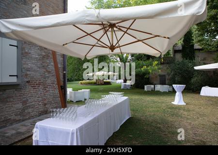 Eleganter langer Tisch mit weißer Tischdecke und vielen leeren Sektgläsern unter einem großen weißen Regenschirm, vorbereitet für eine Hochzeitsfeier in einem Griee Stockfoto