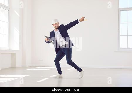 Glücklicher, lustiger Architekt, der Helm und Anzug trägt und in leerer Wohnung steht und Spaß hat. Stockfoto