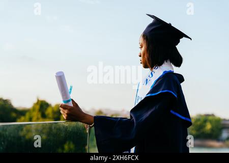 Porträt einer traurigen Studentin an ihrem Abschlussfeiertag auf dem Dach mit Blick auf den Sonnenuntergang auf der Stadt im Hintergrund. Stockfoto