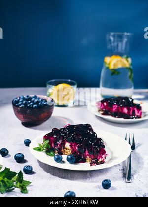Eine Nahaufnahme einer Blaubeeren-Käsekuchenscheibe mit frischen Blaubeeren und einem Glas Zitronenwasser im Hintergrund. Stockfoto