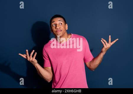 Foto von jungen lustigen Kerl in rosa stilvoll T-Shirt zucken Schultern weiß es nicht leid kann Ihnen jetzt auf dunkelblauem Hintergrund helfen Stockfoto