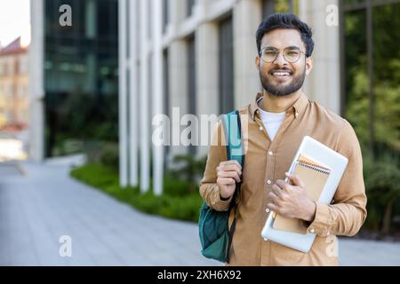 Selbstbewusster Student, der vor dem modernen Universitätsgebäude steht. Junger Mann mit Brille, Rucksack, Büchern und Laptop. Das Konzept der Bildung, des College-Lebens, des akademischen Erfolgs, des Studiums Stockfoto