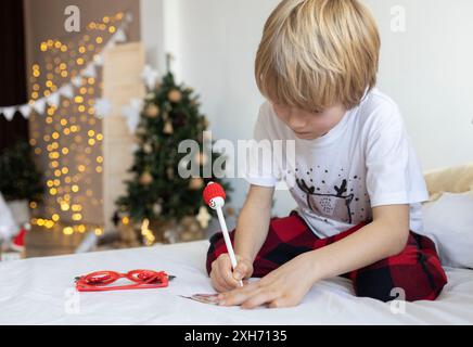 Der kleine Junge schreibt einen Brief an den Weihnachtsmann und träumt von einem Geschenk vor dem Hintergrund von Dekorationen und Lichtern in Innenräumen. Frohe Weihnachten und Hap Stockfoto