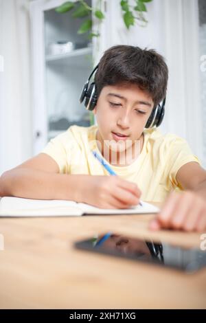Ein Teenager mit Kopfhörern schreibt im Wohnzimmer zu Hause auf Notizblock mit Stift. Über dem Schreibtisch ist das Handy des Jungen. Stockfoto