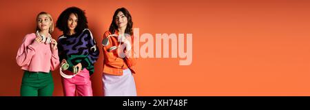 Drei junge, schöne Frauen mit Kopfhörern, die vor einer markanten orangefarbenen Wand posieren. Stockfoto