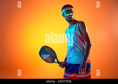 Porträt eines Mannes in Sonnenbrille und Sportbekleidung, stehend mit Tennisschläger am Strand und zeigt Motivation für den Sieg in einem Spiel gegen das orange Studio Stockfoto