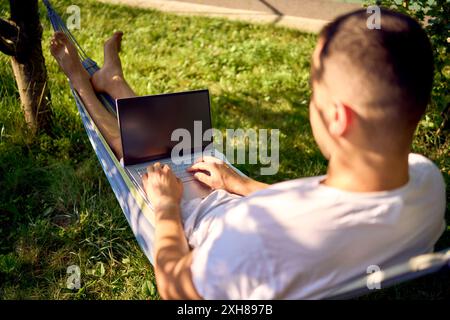 Ein Mann arbeitet an einem Laptop in einer Hängematte im Garten, neben seinem Hund Stockfoto