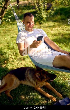 Ein Mann ruht sich aus und hört Musik in einer Hängematte, sein Hund ruht in der Nähe Stockfoto