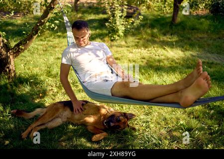 Ein Mann ruht sich aus und hört Musik in einer Hängematte, sein Hund ruht in der Nähe Stockfoto