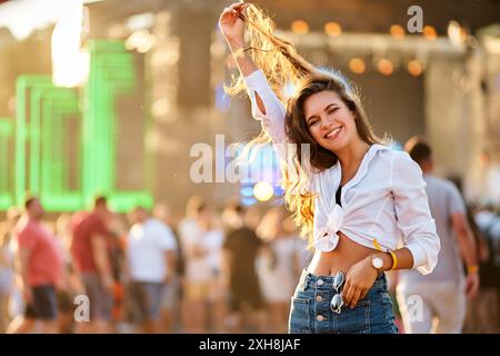 Glückliche junge Frau genießt Sommermusikfestival am Strand. Lächelnde Frau in lässigem Outfit tanzt, Sonnenlicht fackelt dahinter. Die Menge trifft sich im Freien Stockfoto