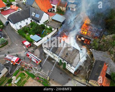 Seesbach, Deutschland. Juli 2024. Feuerwehrleute versuchen, eine brennende Scheune zu löschen. Seesbach (dpa/lrs) Eine Scheune brennt in der Innenstadt und hat zwei Wohngebäude getroffen. Das Feuer habe sich auf den Dachstuhl eines angrenzenden Hauses ausgebreitet, das wegen Renovierungsarbeiten leer war, sagte ein Polizeisprecher. Die Fassade des zweiten Gebäudes war betroffen. Soweit bekannt, wurde niemand verletzt. Die Löscharbeiten sind noch nicht abgeschlossen, sagte der Sprecher. (Aufgenommen mit einer Drohne) Credit: Sebastian Schmitt/dpa/Alamy Live News Stockfoto