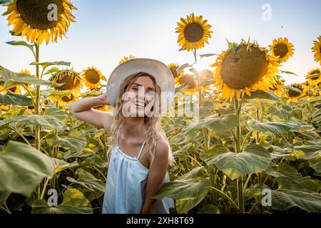 Eine Frau mit langen blonden Haaren lächelt, während sie einen großen weißen Hut in einem Feld voller Sonnenblumen trägt. Stockfoto