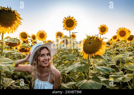 Eine Frau in weißem Kleid und Hut lächelt, während sie in einem blühenden Sonnenblumenfeld posiert. Stockfoto