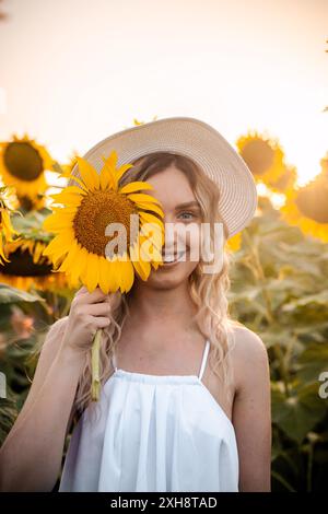 Eine Frau mit langen, blonden Haaren trägt ein weißes Tanktop und einen Strohhut. Sie hält eine große Sonnenblume vor ihrem Gesicht und lächelt in einem Sonnenblumenfeld. Stockfoto