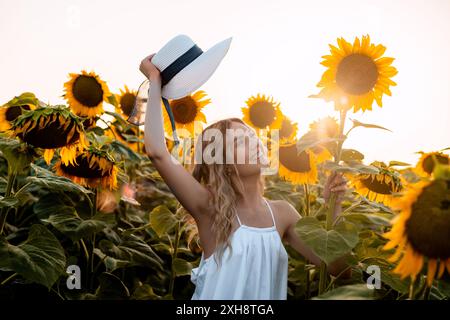 Eine Frau mit langen blonden Haaren steht in einem Sonnenblumenfeld mit erhobener Hand und einem Strohhut. Die Frau trägt ein weißes Sommerkleid und Stockfoto