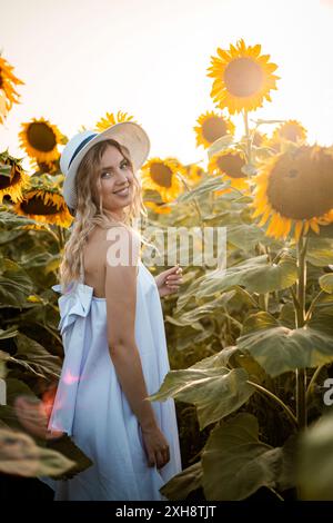 Eine Frau in einem weißen Kleid lächelt in die Kamera, während sie in einem Feld voller Sonnenblumen steht. Sie trägt einen Strohhut und die Sonne untergeht im Hintergrund Stockfoto