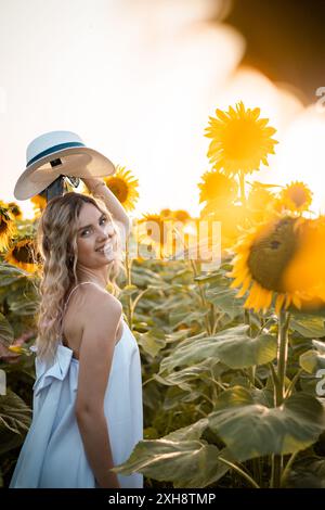 Eine Frau in einem weißen Kleid lächelt in einem Feld voller Sonnenblumen, während sie einen breitkrempigen Hut hält. Stockfoto