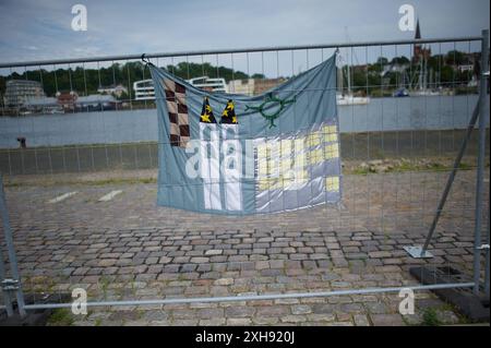 Flensburg, Schleswig-Holstein Dampfrundum 2024 Flensburg, transparent am Bauzaun. Das Dampfrundum 2024 wird aufgrund der Schäden durch das Ostseesturmhochwasser im Oktober 2023 an einem anderen Ort stattfinden. Die Organisatoren müssen auf den angestammten Platz an der südlichen Schiffbrücke verzichten, da die Kaikante dort abgesackt ist. Eine Absperrung wurde errichtet, hier: Blick von der Westseite des Flensburger Hafens zur Ostseite über das Hafenbecken. Aufnahme vom 12.07.2024, Flensburg, Hafen West *** Flensburg, Schleswig-Holstein Dampfrundum 2024 Flensburg, Banner auf dem Bauofen Stockfoto
