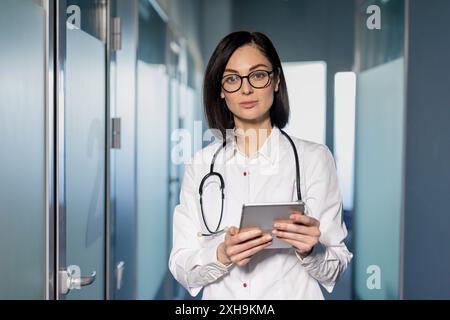 Selbstbewusste Ärztin mit Brille und Stethoskop mit digitalem Tablet im modernen Krankenhausflur. Professionelle medizinische Fachkraft mit Technologie, bereit, Patienten zu helfen. Stockfoto