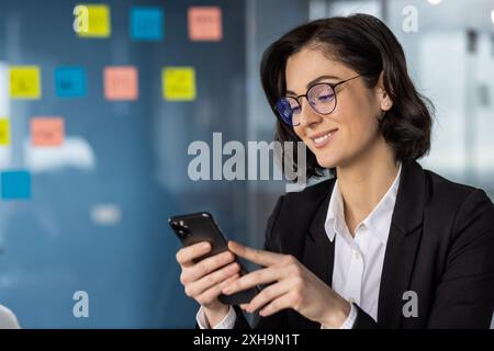 Selbstbewusste Geschäftsfrau mit Brille und Smartphone im modernen Büro. Durchdachte professionelle Überprüfung von Nachrichten auf dem Telefon, umgeben von Haftnotizen. Abendliche Arbeitsatmosphäre Stockfoto