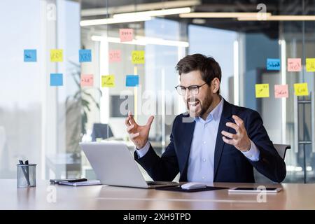 Wütender Mann im Business-Anzug, der im Büro den Laptop anschreit, reifer Geschäftsmann-Chef am Arbeitsplatz, der mit der Leistung des Unternehmens unzufrieden ist, streitet mit Stockfoto