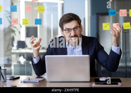 Wütender Chef im Büro am Arbeitsplatz, erfahrener Geschäftsmann, der die Kamera schreien sieht, älterer reifer Mann im Business-Anzug, der mit einem Laptop arbeitet. Stockfoto
