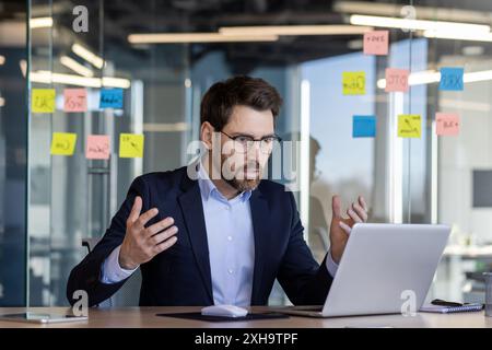 Wütender Mann im Business-Anzug, der im Büro den Laptop anschreit, reifer Geschäftsmann-Chef am Arbeitsplatz, der mit der Leistung des Unternehmens unzufrieden ist, streitet mit Stockfoto