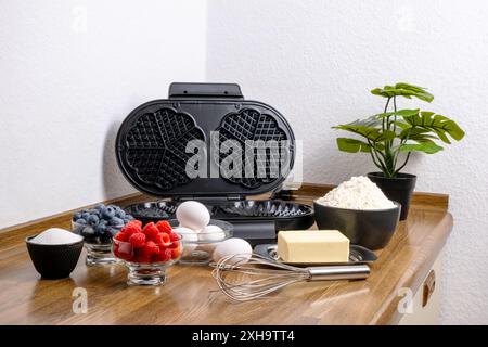 Zutaten für belgische Waffeln und Waffeleisen auf dem Küchentisch. Eier, Butter, Zucker, Mehl und Schneebesen zur Herstellung von Teig und Beeren. Waffeln machen Stockfoto