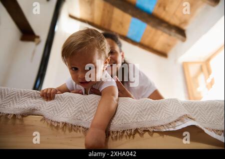 Ein fröhliches Kleinkind, das mit seiner Mutter auf einem Holzbett in einem hellen und gemütlichen Zuhause spielt, schafft einen warmen und liebevollen Familienmoment. Stockfoto