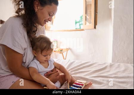 Eine glückliche Mutter genießt die schöne Zeit mit ihrem Baby, während sie an einem sonnigen Tag drinnen mit einem farbenfrohen Xylophon spielt und eine warme und fröhliche Atmosphäre schafft. Stockfoto