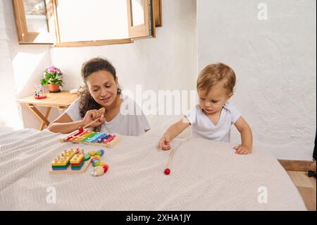 Mutter und Kleinkind spielen mit bunten Lernspielzeugen auf einem Bett in einem hellen Raum mit Sonnenlicht, das durch das Fenster strömt. Stockfoto