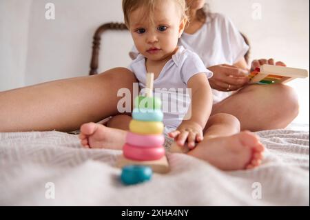 Ein Baby spielt auf einem Bett mit einem pädagogischen Spielzeug, während die Mutter im Hintergrund ist, und betont spielerisches Lernen und Entwicklungswachstum. Stockfoto