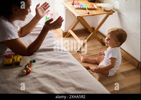 Eine Mutter, die fröhlich mit ihrem kleinen Kind zu Hause spielt. Das Kind sitzt auf dem Boden, umgeben von farbenfrohen Spielzeugen, die eine warme und liebevolle Atmosphäre schaffen Stockfoto