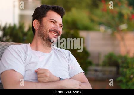 Einsamer, fröhlich lächelnder bärtiger Mann mittleren Alters sitzt draußen bei Sonnenuntergang. Happy Life-Konzept Stockfoto