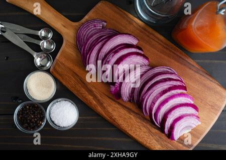 Zutaten für eingelegte Zwiebeln vorbereiten: Geschnittene rote Zwiebeln auf einem hölzernen Schneidebrett mit koscherem Verkauf, schwarze Pfeffer, Zucker und Essigflaschen Stockfoto