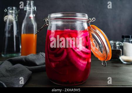 Glas mit eingelegten Zwiebeln von der Seite aus gesehen: Hell gefärbte geschnittene rote Zwiebeln, eingelegt in einem einmauerglas, umgeben von Zutaten Stockfoto