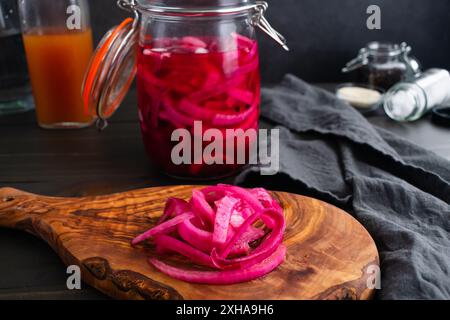 Nahansicht eingelegter Zwiebeln auf einem Schneidebrett: In hellen Farben geschnittene rote Zwiebeln, eingelegt in einem einmauerglas, umgeben von Zutaten Stockfoto