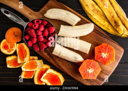 Frisches Obst für eine Smoothie Bowl vorbereiten: Himbeeren mit geschälten und halbierten Bananen und cara cara Orange auf einem hölzernen Schneidebrett Stockfoto