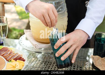 Luxuriöses Catering bei Gartenparty im Sommer. Die Hände des Kellners gießen erfrischende Limonade aus dem Glas in ein schickes Glas. Gäste, die den eleganten Außenbereich genießen Stockfoto