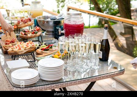 Ein langer Tisch mit Speisen und Getränken für das Catering im Freien. Enthält Sektflöten, Obstpunsch-Spender, Vorspeisen. In wunderschönem Outdoor-Set Stockfoto