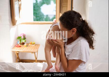 Die liebevolle Mutter hält ihr Baby sanft in einem hellen, gemütlichen Raum, der mit natürlichem Sonnenlicht gefüllt ist, und küsst es. Schöner Moment der mütterlichen Pflege und Zuneigung in Stockfoto